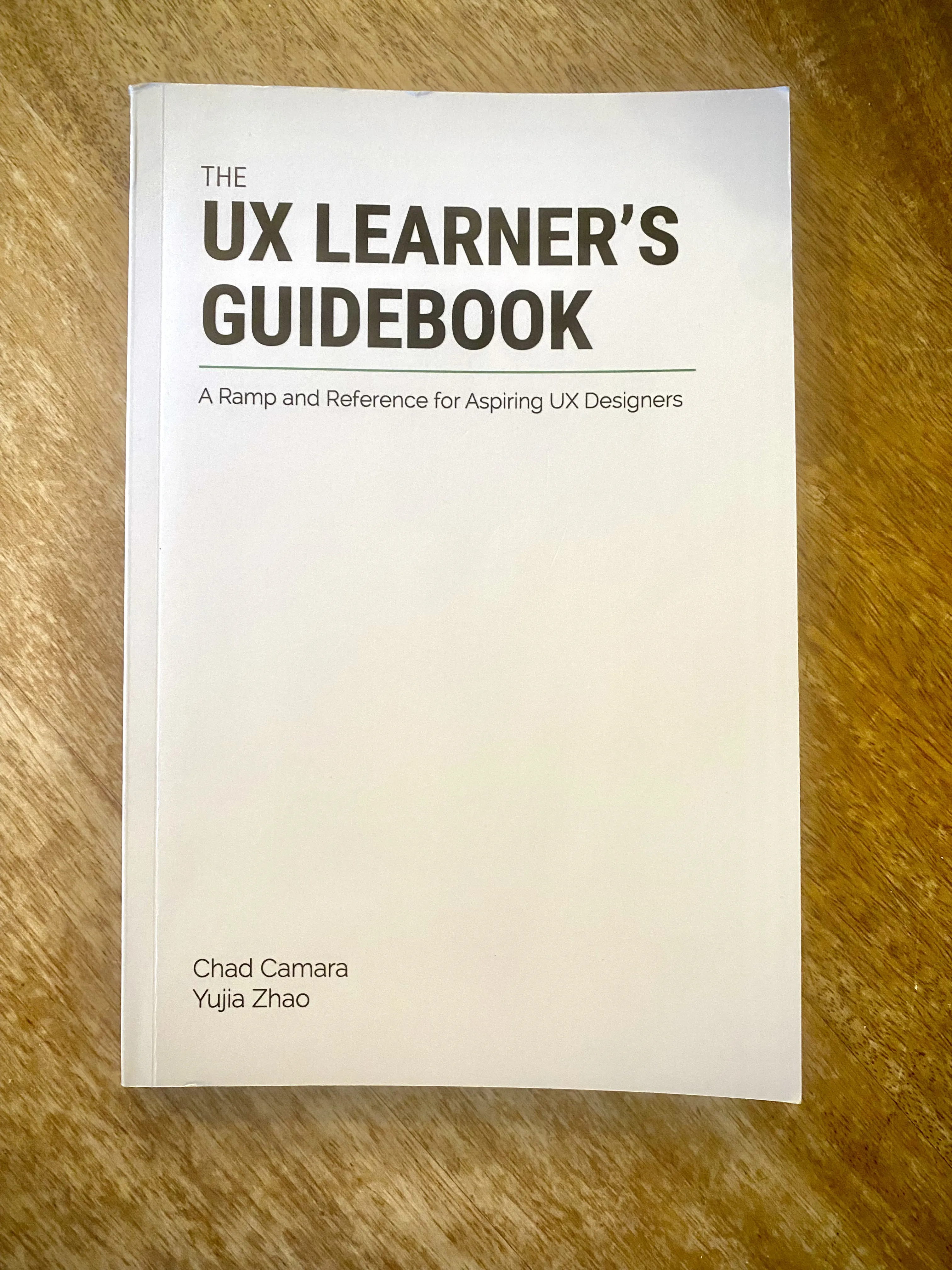 The printed paperback of The UX Learner's Guidebook, subtitled "A Ramp and Reference for Aspiring UX Designers", by Chad Camara and Yujia Zhao, lying on a wooden table.